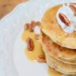 A stack of Cracker Barrel Pecan Pancakes topped with pecans and whipped cream with a drizzle of maple syrup.